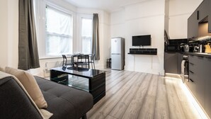 A modern living room with a glass coffee table, a sofa, and a dining area with chairs. A kitchen with a stove, microwave, and cabinets is visible in the background.