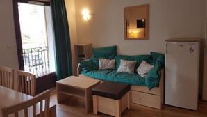 A small, cozy room with a green sofa, a wooden table, a bench, a small cabinet, and a refrigerator. - Lovely apartment in LUCHON, in a secure residence, wooded grounds (BAGNERES DE LUCHON)