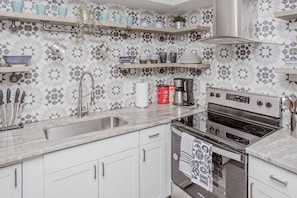 Una cocina con armarios blancos, un lavamanos de acero inoxidable, una estufa y repisas con utensilios de cocina.
