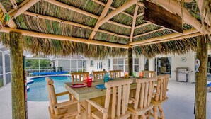 Una mesa de comedor de madera con sillas bajo un techo de paja, una piscina y vistas a una casa.