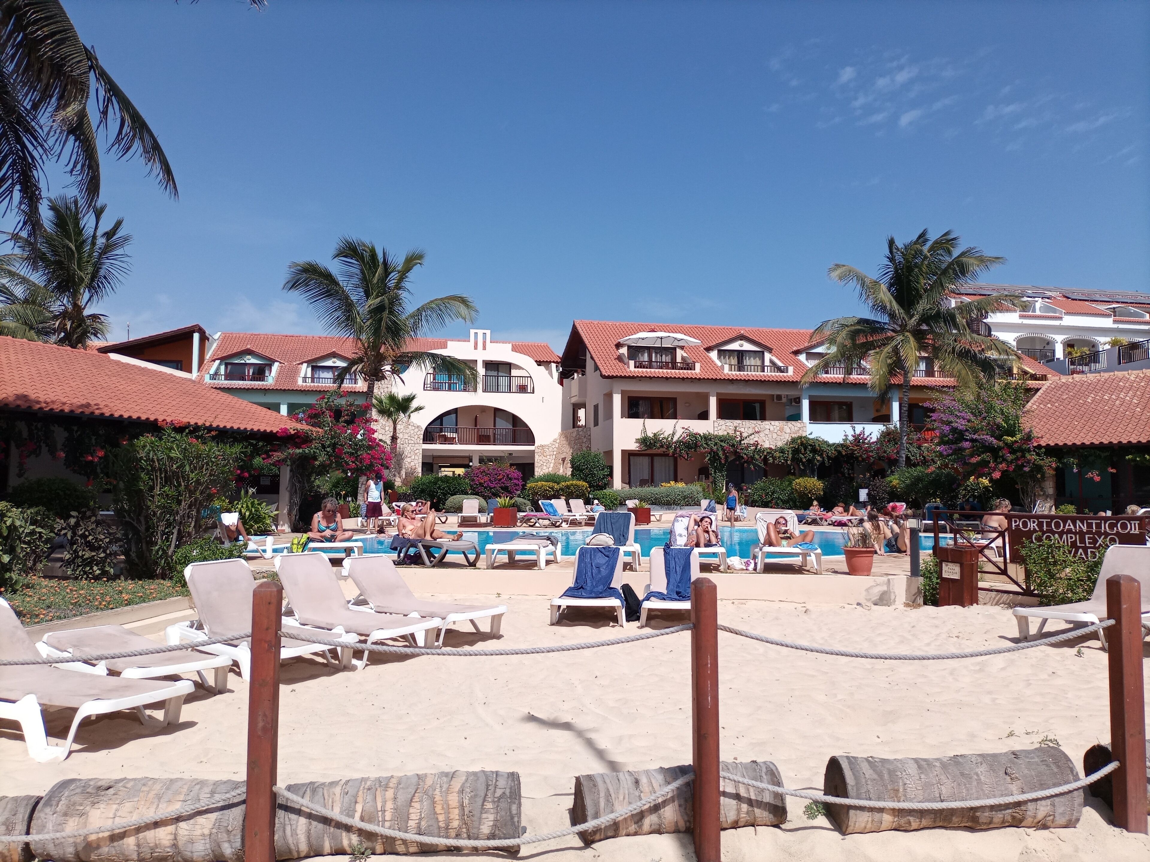 Un resort con varias construcciones, un área de piscina con sillones de descanso y una playa de arena.