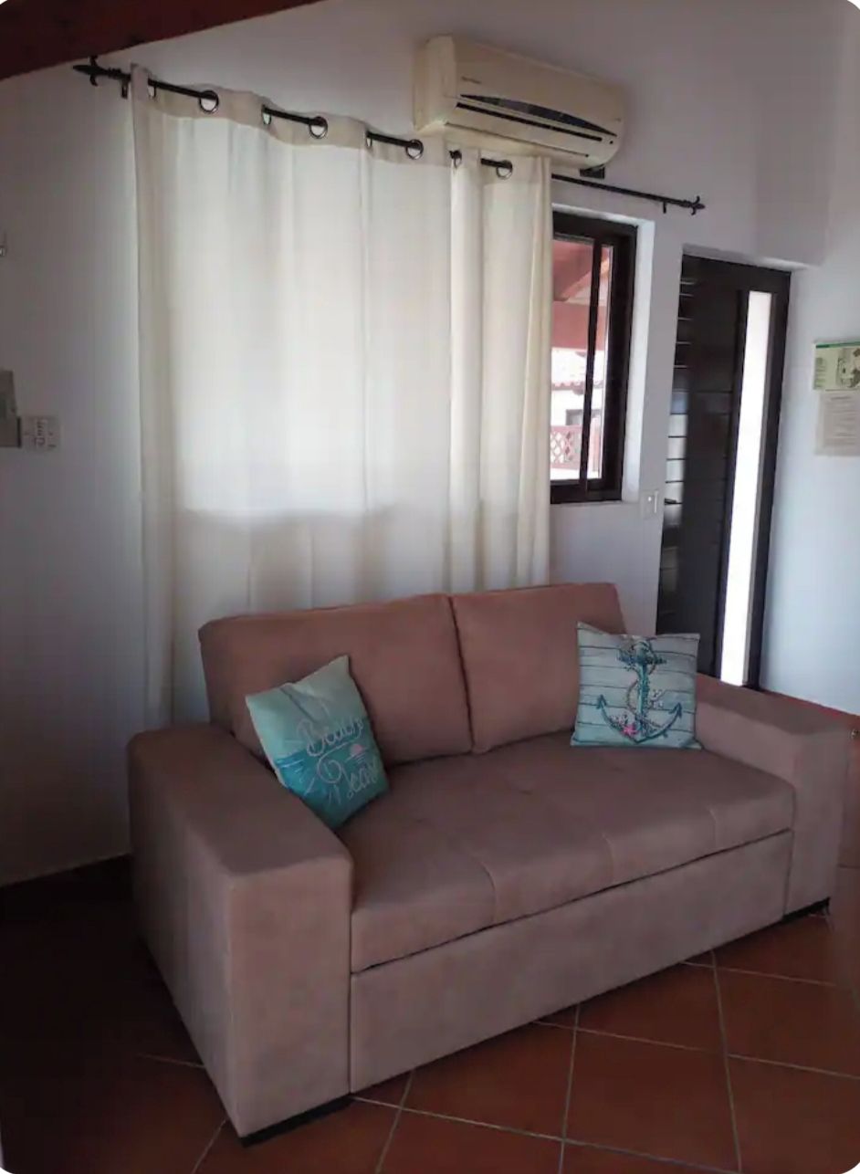 A living room with a beige sofa, two cushions, a window with curtains, and an air conditioning unit.