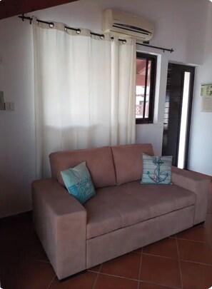 A living room with a beige sofa, two cushions, a window with curtains, and an air conditioning unit.