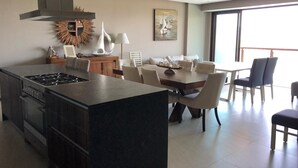 A modern kitchen with a central island, dining area with chairs, and a large window providing natural light. - Luxury apartment in Acapulco diamond (Acapulco de Juárez)