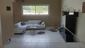 A living room with a white sofa, a small table, a TV, and a window with a view of greenery. - Villa with private pool and jacuzzi, lovely view (Herry)