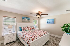 A bedroom with a bed, nightstand, lamp, ceiling fan, and two paintings on the wall.