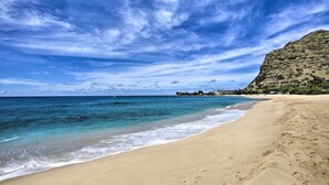 A beach with clear blue water, a sandy shore, and a rocky hill in the background. - King size Bed, 1BR condo, Makaha Valley, with Mtn and Ocean views (Waianae)