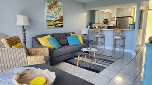 A modern living room with a grey sofa, yellow and blue cushions, a woven chair, a small round table, and a kitchen area with a fridge and bar stools. - King size Bed, 1BR condo, Makaha Valley, with Mtn and Ocean views (Waianae)