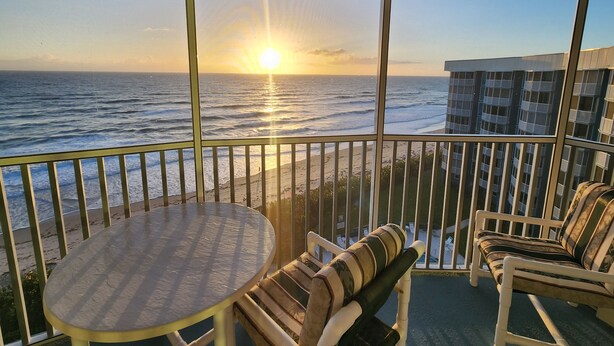 Ein Balkon mit Blick auf das Meer bei Sonnenuntergang, ein runder Tisch und zwei Stühle mit gestreiften Kissen.