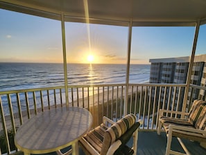 Un balcon offrant une vue sur l’océan au coucher du soleil, une table ronde et deux chaises avec des coussins à rayures.