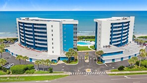 A beachfront hotel with two blue and white buildings, a swimming pool, and a parking area.