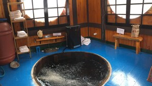 A room with a large black bathtub, wooden shelves with towels, a wooden bench, and a sign indicating the bathtub is for use.