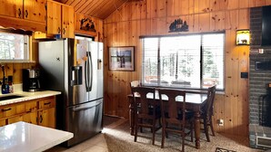 A wooden cabin interior with a kitchen area, a dining table with chairs, a refrigerator, and a fireplace. - Cabin at Sportsman’s Paradise- Anglers’s Dream & Mountain Retreat. (Lake George)