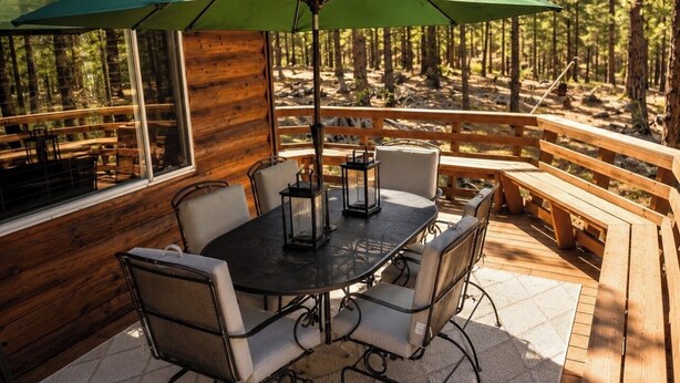 A wooden deck with a table and chairs, a green umbrella, and a view of a forest. - Cabin at Sportsman’s Paradise- Anglers’s Dream & Mountain Retreat. (Lake George)