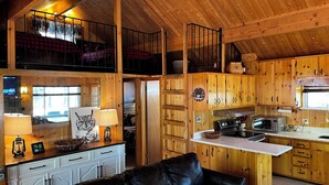 A cozy cabin interior with a dining area, kitchen, and a loft bedroom with a balcony. - Cabin at Sportsman’s Paradise- Anglers’s Dream & Mountain Retreat. (Lake George)