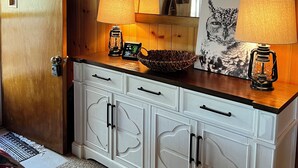 A cabin interior with wooden walls, a large mirror, a white cabinet with drawers, a woven basket, and two lanterns. - Cabin at Sportsman’s Paradise- Anglers’s Dream & Mountain Retreat. (Lake George)