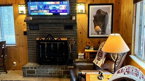 A cozy living room with a fireplace, a flat-screen TV mounted on the wall, and a comfortable leather sofa. - Cabin at Sportsman’s Paradise- Anglers’s Dream & Mountain Retreat. (Lake George)