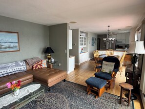 Uma sala de estar com um sofá de couro marrom, uma mesa de centro de vidro com um sousplat rendado, um tapete estampado e uma área de jantar com cadeiras de madeira e uma mesa.
