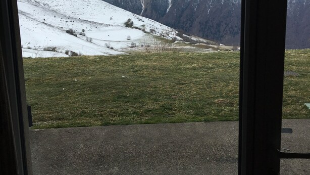 Vista a través de una ventana de un paisaje montañoso con picos nevados y un primer plano con césped.