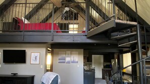Un vaste espace de vie doté d’un haut plafond, d’un ventilateur de plafond en bois, d’un fauteuil bleu, d’un escalier menant à un niveau supérieur et d’un coin cuisine à l’arrière-plan.