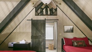 Une chambre avec un plafond en pente, une porte en bois, un lit avec une literie rouge, une table de chevet et un petit tableau accroché au mur.