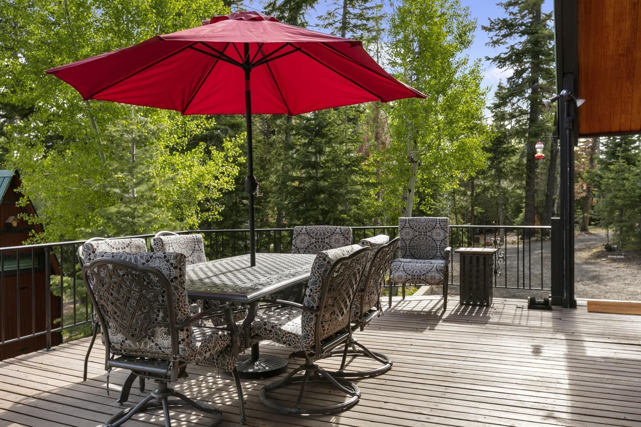 A wooden deck with a table and chairs, a red umbrella, and a view of trees.
