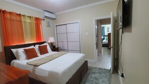 Een slaapkamer met een bed, nachtkastje, airconditioning, televisie en een deur naar een andere kamer.