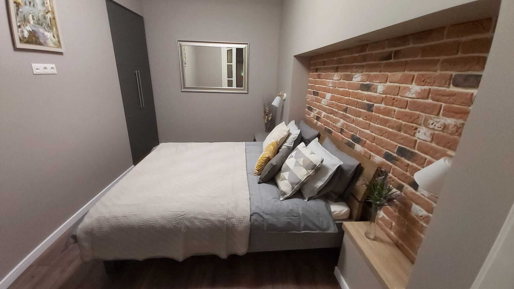 Une chambre avec un lit, un miroir, un mur en briques et une petite fenêtre.