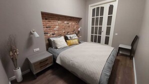 Un dormitorio con cabecera de ladrillo, cama con cubrecama gris y blanco, mesita de noche con lámpara y una silla.