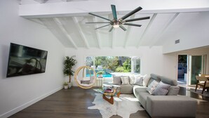 A spacious living room with a large flat-screen TV, a ceiling fan, a hanging chair, a glass coffee table, and a view of a backyard with a pool. - Amazing Views with Heated Pool and Jacuzzi Near Beach (Solana Beach)