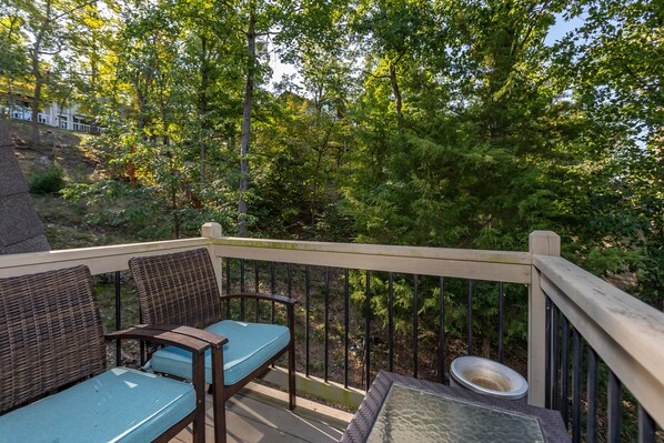 A wooden deck with wicker furniture and a glass table, surrounded by a metal railing and dense greenery.