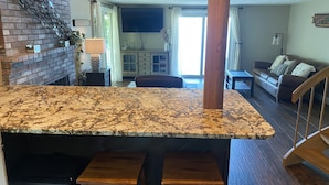 Una cocina con encimera de granito, taburetes de madera, pared de ladrillo y vistas al salón.