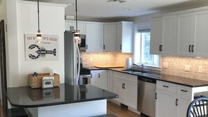 Cocina moderna con armarios blancos, encimera negra y suelo de madera. Hay tres taburetes negros y una lámpara colgante.