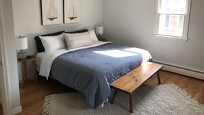 Un dormitorio con cama, una banca, una mesita de noche, una lámpara y dos cuadros de veleros en la pared.