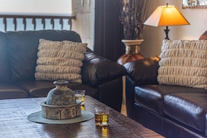 A cozy living room with a leather sofa, a small table with a decorative vase and glasses of whiskey, and a lamp providing warm lighting.
