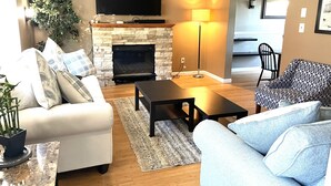 A living room with a fireplace, a flat-screen TV mounted above it, a sofa, a coffee table, and a patterned armchair.