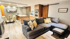 Een moderne woonkamer met een grijze hoekbank, een eethoek met een tafel en stoelen, en een keuken met een magnetron en oven.