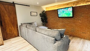 Un salon moderne avec un canapé d’angle gris, un téléviseur fixé au mur, une porte coulissante en bois et un mur en briques.