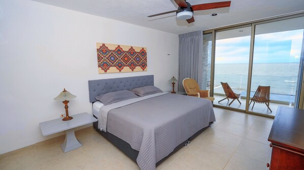 A bedroom with a bed, a ceiling fan, a nightstand, a bench, a chair, and a view of the ocean through large windows.
