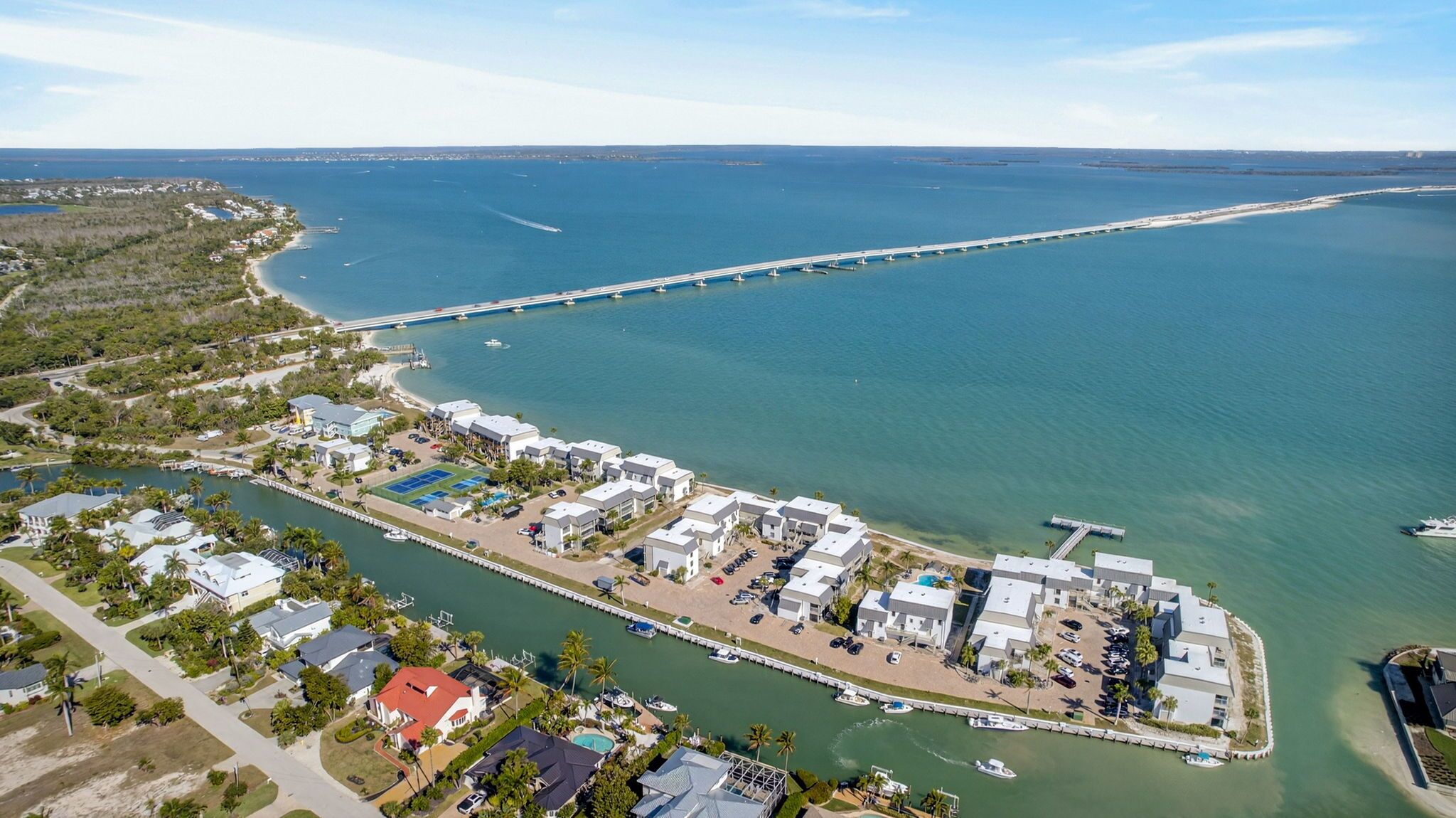 An aerial view of a coastal area with a marina, a long pier, and a row of buildings with parking and swimming pools.