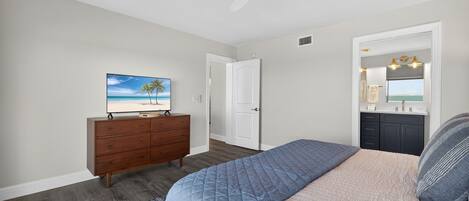 Un dormitorio con una cama, un aparador, un ventilador de techo, un cuadro de una playa y un baño con espejo y lavabo.