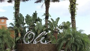 Uma área ajardinada com uma grande placa de metal que diz "Ole", rodeada por palmeiras e um edifício com torre do relógio ao fundo.