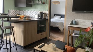 A modern kitchen with a dining area, a small table with a remote control, and a bed in the adjacent room. - Le Clos des Feugrais*** - Quiet outbuilding (Saint-Aubin-lès-Elbeuf)