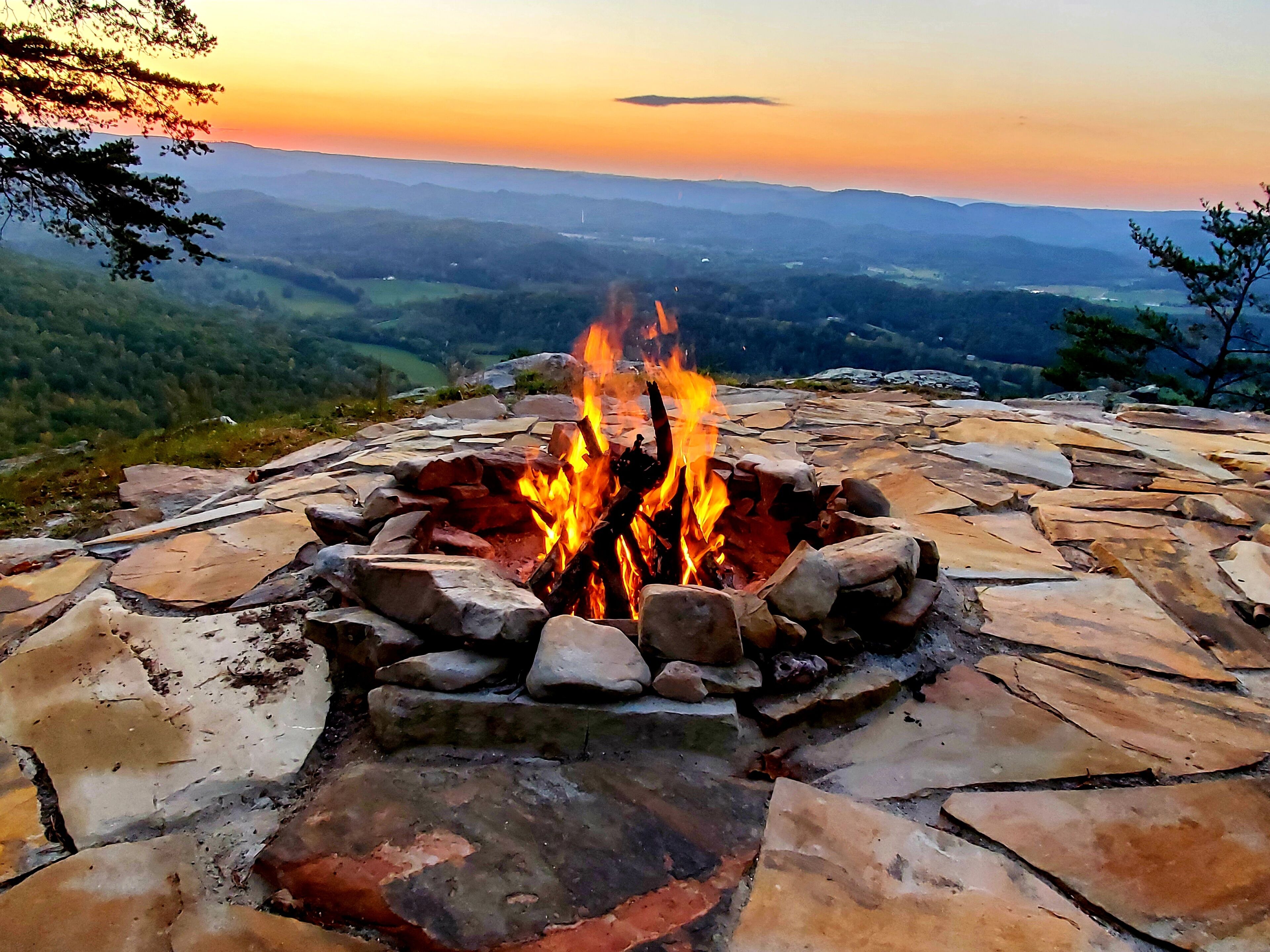 A fire pit surrounded by stone slabs, set against a backdrop of a mountainous landscape during sunset.