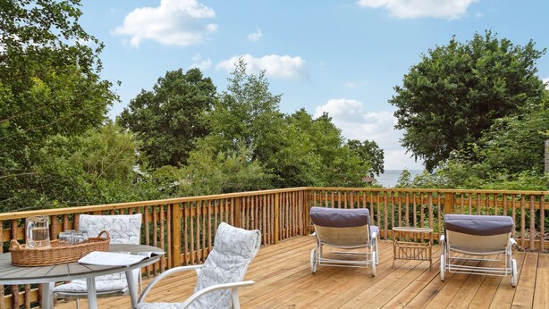 Une terrasse en bois, agréée de mobilier d’extérieur, notamment des chaises et une table, qui offre une vue sur une étendue d’eau, avec des arbres en arrière-plan.
