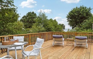 Eine Holzterrasse mit Gartenmöbeln, darunter Stühle und ein Tisch, mit Blick auf ein Gewässer, im Hintergrund Bäume.