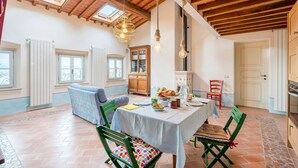 Un coin repas avec une table dressée pour quatre personnes, entouré de chaises vertes agrémentées de coussins colorés. On y trouve deux fenêtres à cadre blanc, un meuble en bois et un plafond avec des poutres apparentes.