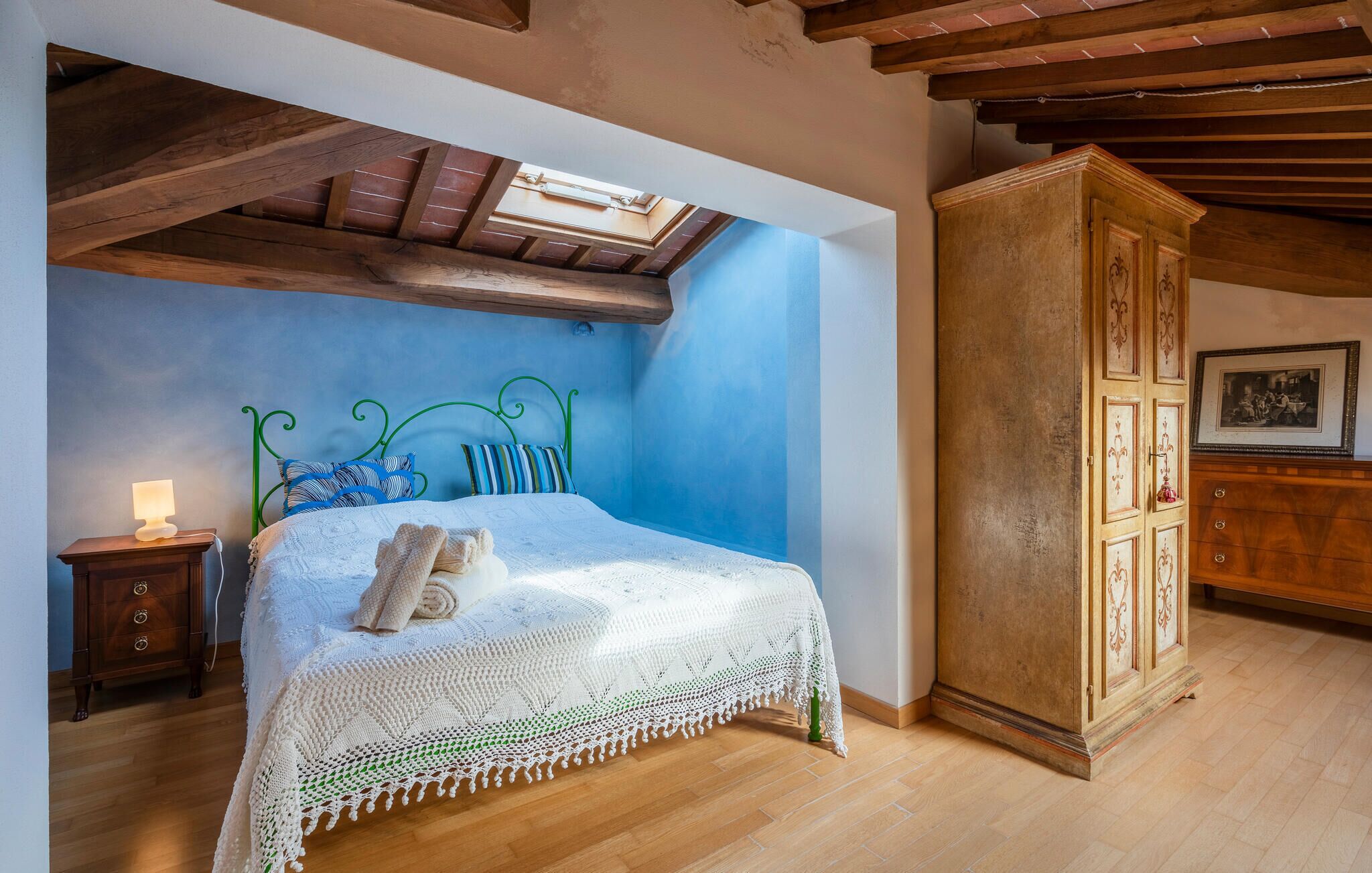 Een slaapkamer met een houten plafond, een bed met een wit gebreide deken, een nachtkastje met een lamp en een houten kast.