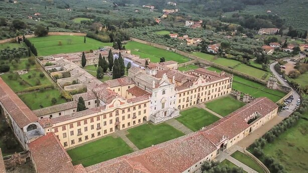 Une vue aérienne d’un grand bâtiment historique à la façade symétrique, entouré de champs verdoyants et de bâtiments plus petits.