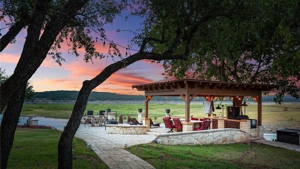 A covered outdoor seating area with stone planters and a grill, overlooking a grassy field and a body of water. - Lake Travis Waterfront LUX/Dock/Firepit/Outdr play (Lago Vista)